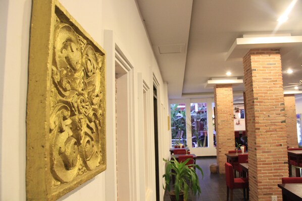 A hallway with a large gold-colored relief sculpture on the wall, brick pillars, and red chairs and tables in the dining area.