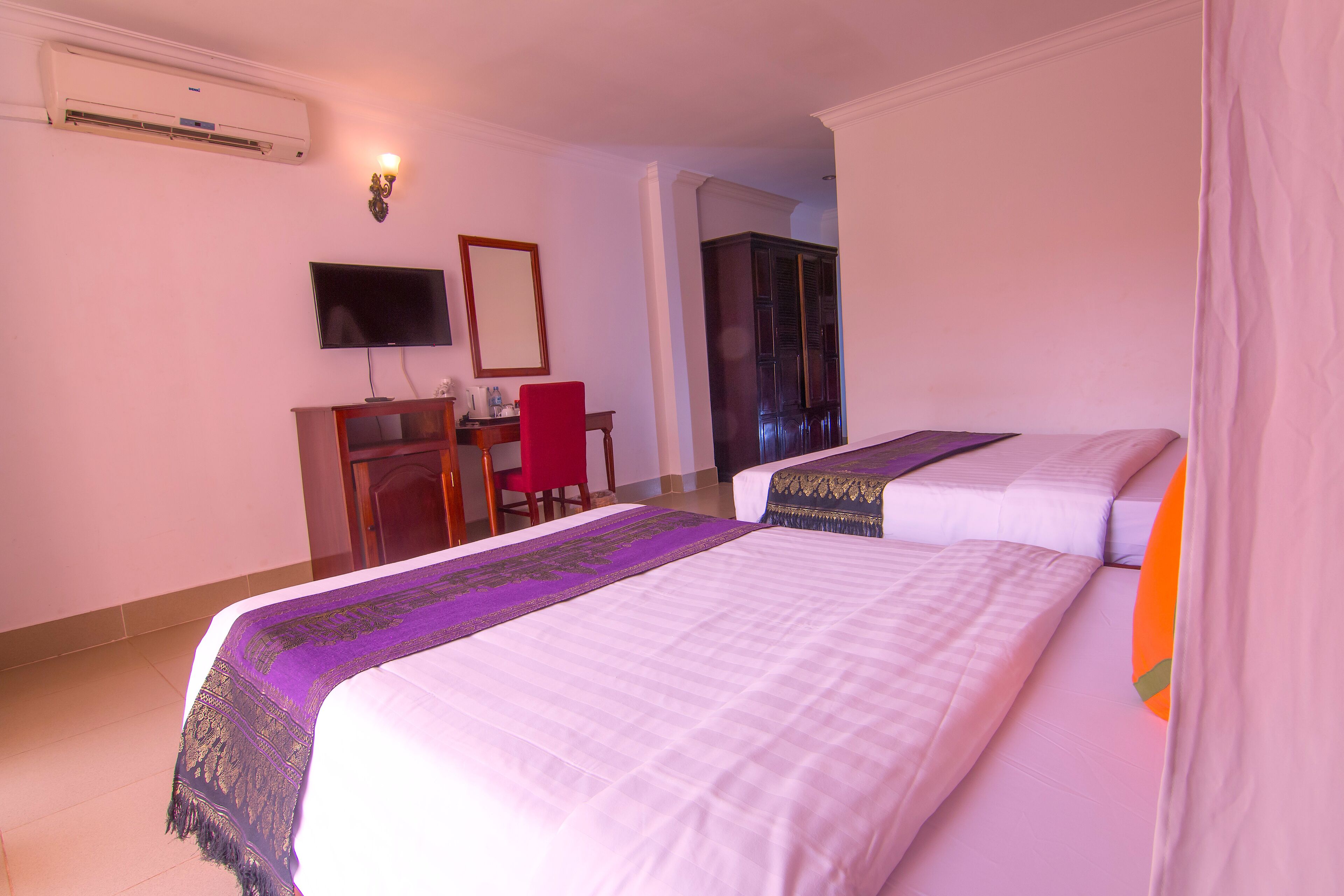 A hotel room with two beds, a desk with a chair, a television, and an air conditioning unit.