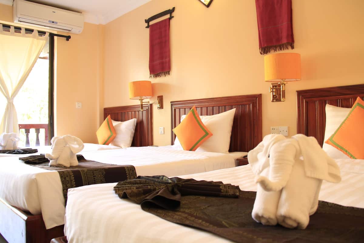 Une chambre d’hôtel avec deux lits, chacun doté d’une tête de lit en bois, de draps blancs et d’oreillers orange. On y trouve une fenêtre avec un rideau, un appareil de climatisation placé au-dessus, et une lampe fixée au mur.