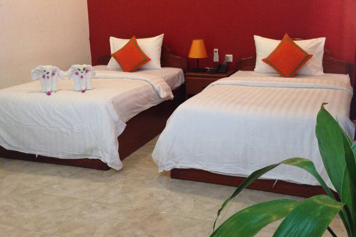 Deux lits simples avec du linge de lit blanc et des oreillers orange, une table de chevet avec une lampe, et une plante au premier plan.