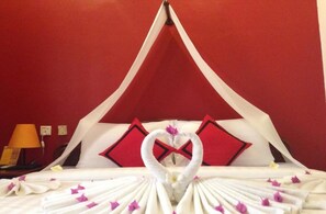A hotel room with a red wall, a canopy bed with white and red bedding, and a heart-shaped towel arrangement.