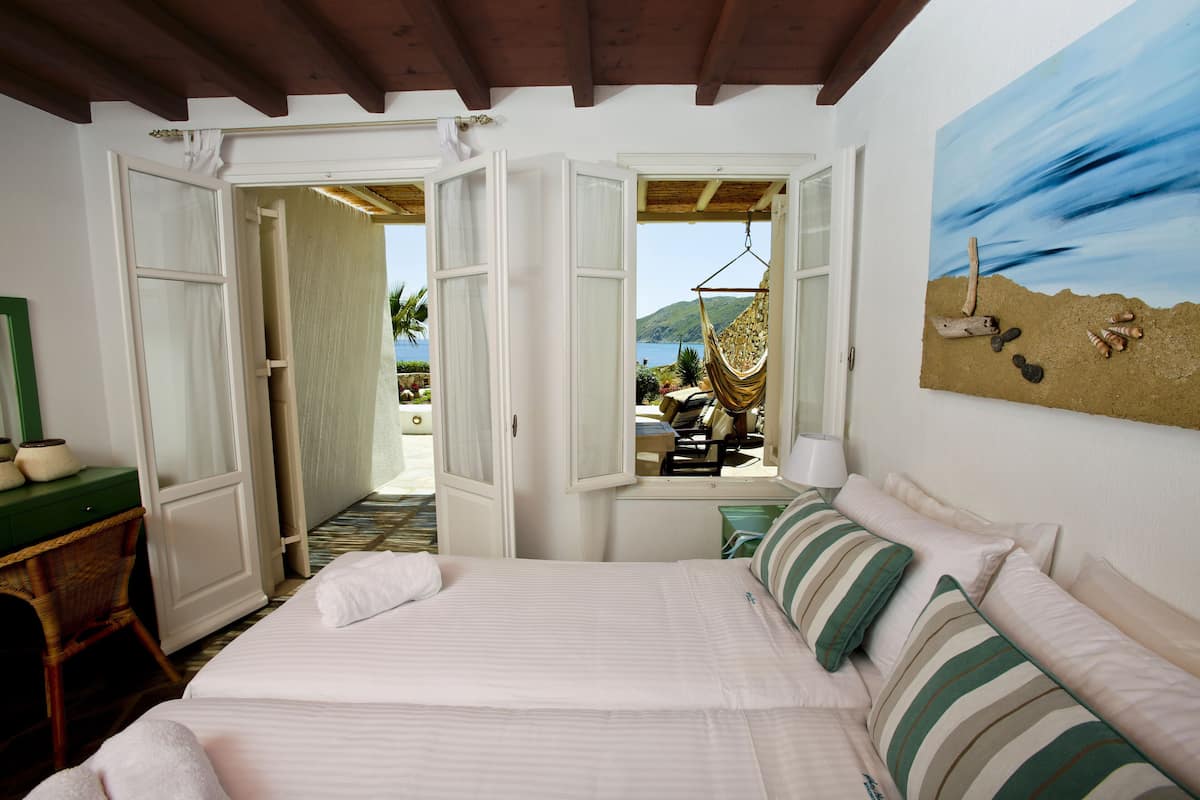 A bedroom with a bed, striped pillows, a painting of a beach scene, and an open door leading to a balcony with a view of the sea.