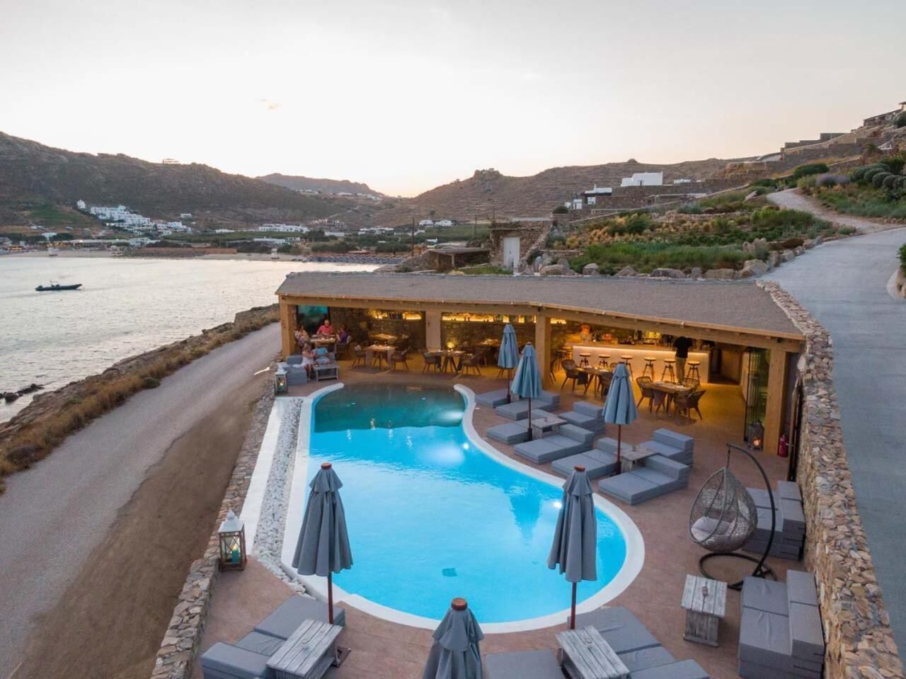 Zona de piscina con sillones de descanso y sombrillas, un bar y vistas al mar y a las colinas.