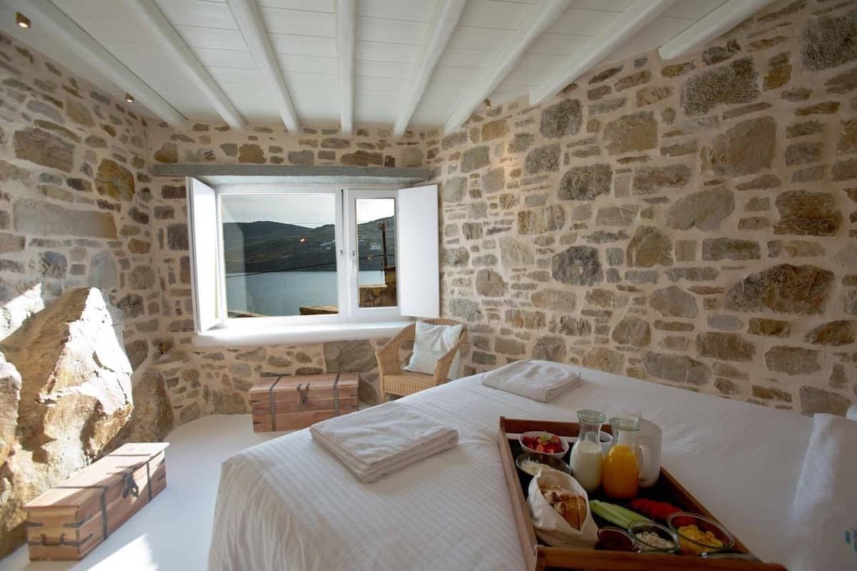 A stone-walled bedroom with a bed, a breakfast tray, and a window overlooking a body of water.