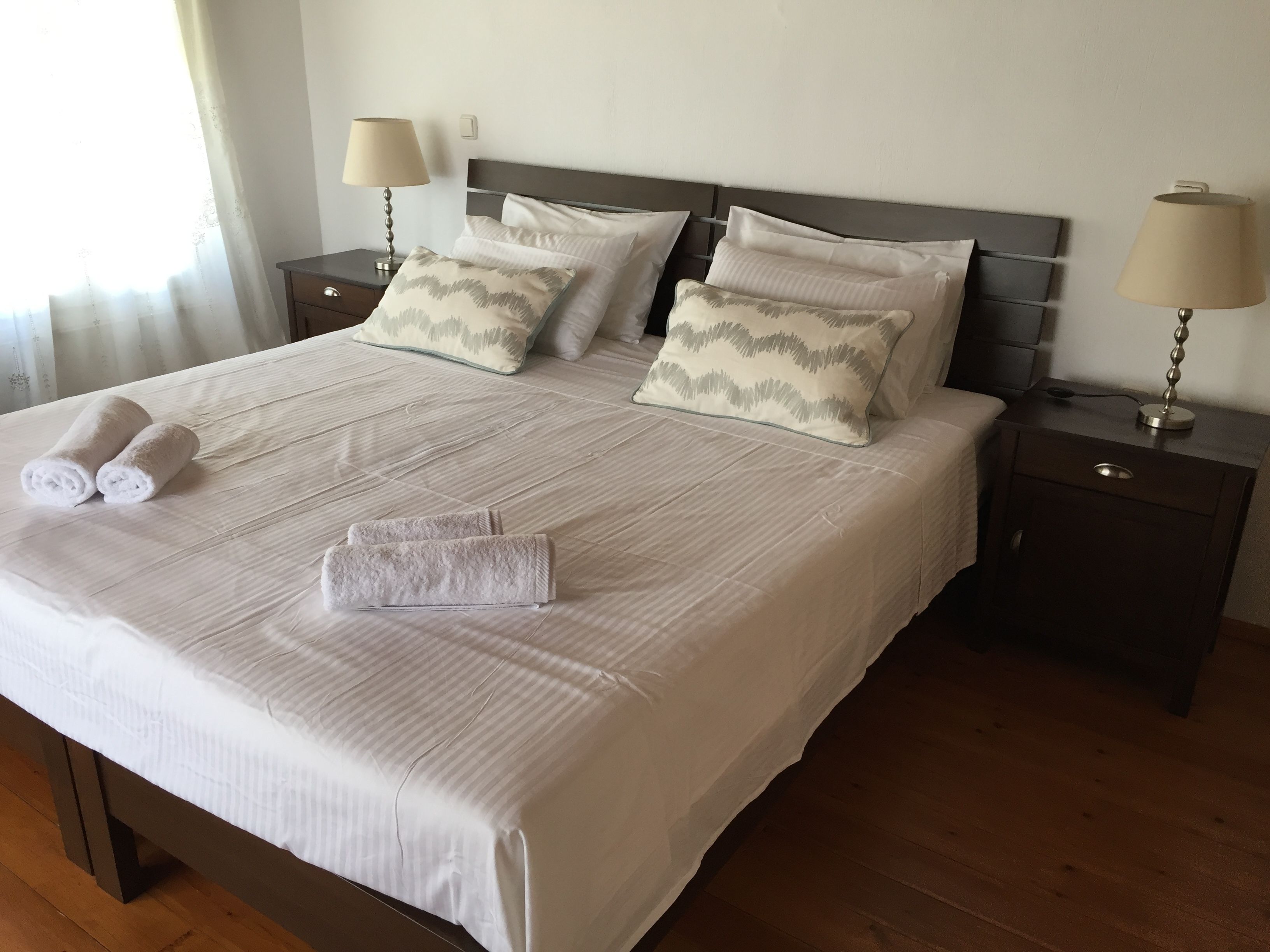A neatly made bed with white linens and two pillows, two bedside lamps, and a wooden nightstand.