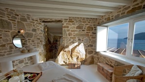 A stone-walled room with a bed, a wicker chair, and a view of the sea. - Salty Houses (Mykonos)
