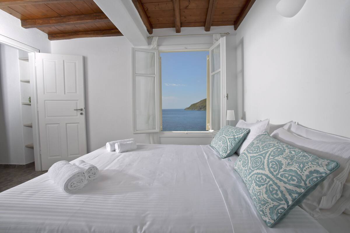 A bedroom with a bed, a window with an open shutter, and a view of the sea.