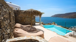 A stone-walled outdoor area with a pool, a thatched-roof structure, and a white building with a dome. - Salty Houses (Mykonos)