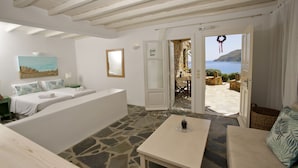 A room with a stone floor, white walls, a bed with white linens, a table with a glass top, and a view of the sea through an open door. - Salty Houses (Mykonos)