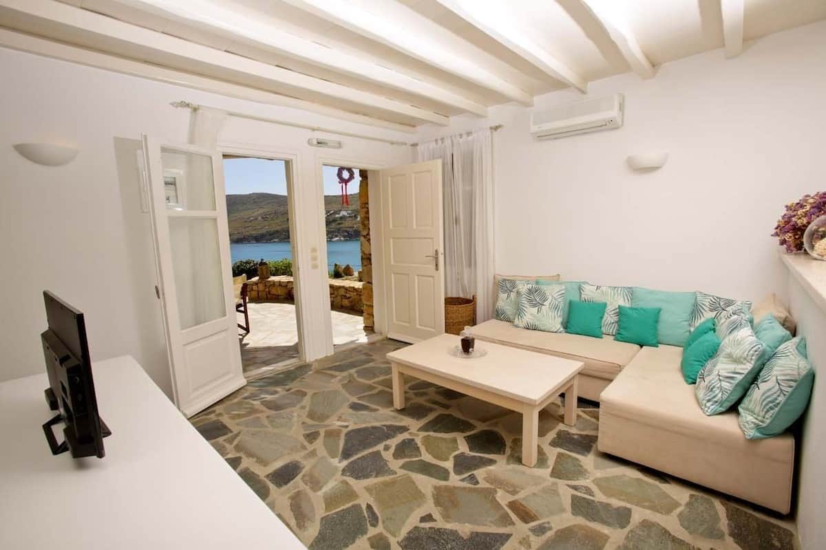 A room with a stone floor, a white sofa with teal cushions, a wooden coffee table, and a television on a white table.