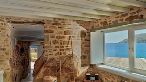 A stone-walled room with a wooden ladder, a window with a view of the sea, and a small table with boxes. - Salty Houses (Mykonos)