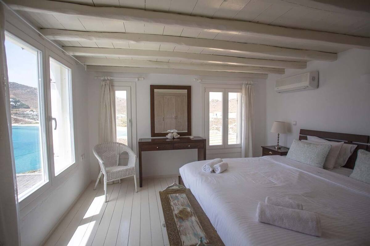 A bedroom with a large bed, a desk, a chair, and a view of the sea.