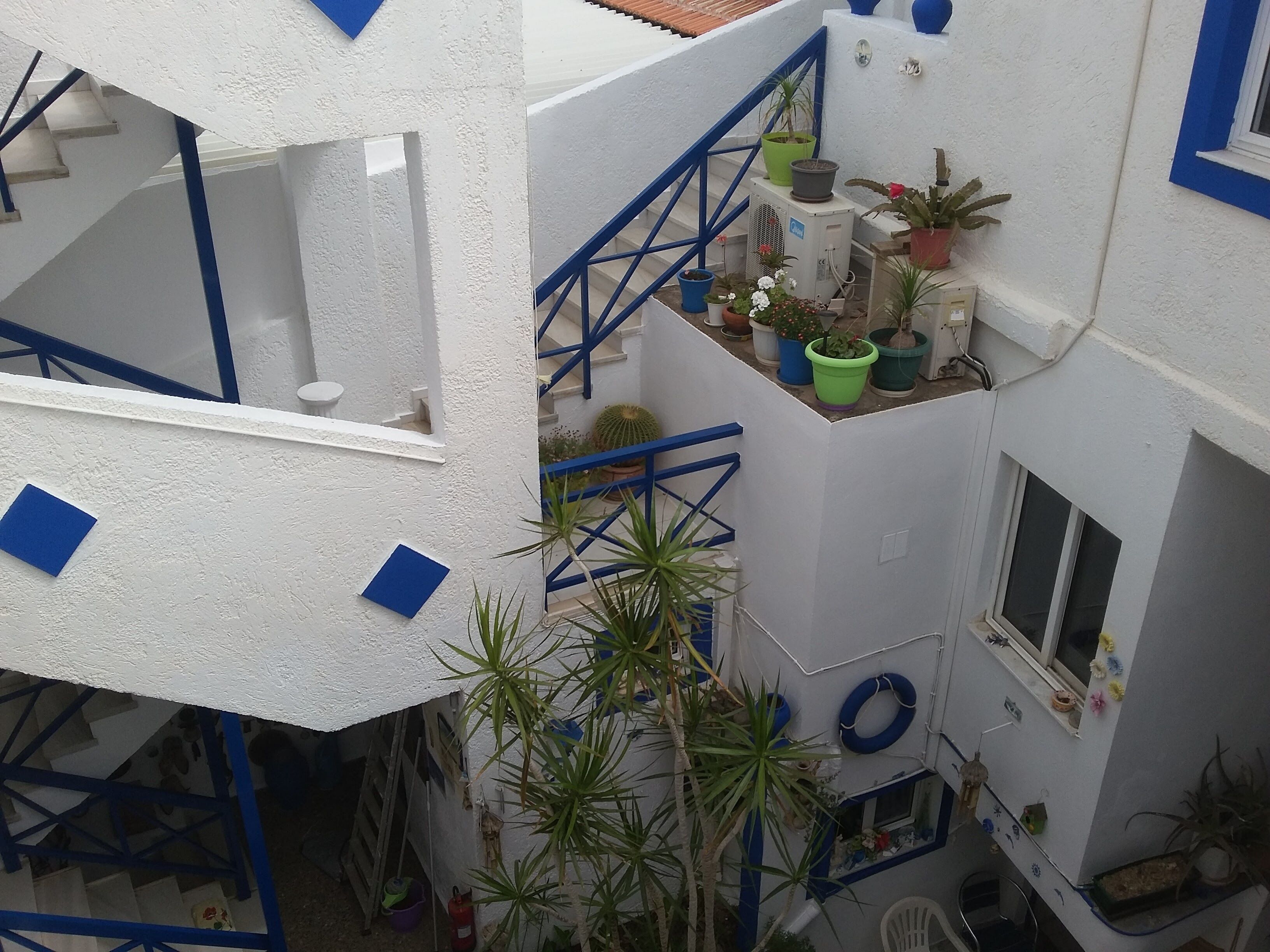 Um edifício residencial com vários andares, corrimãos azuis, paredes brancas e diversos vasos com plantas.