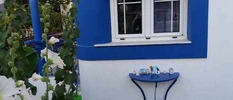 Un bâtiment à l’extérieur bleu et blanc, avec une fenêtre et une petite table bleue sur laquelle se trouvent divers objets.