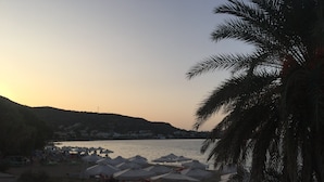 Ein Strand mit Reihen von Sonnenschirmen und Liegestühlen, ein ruhiges Meer und eine hügelige Landschaft im Hintergrund.