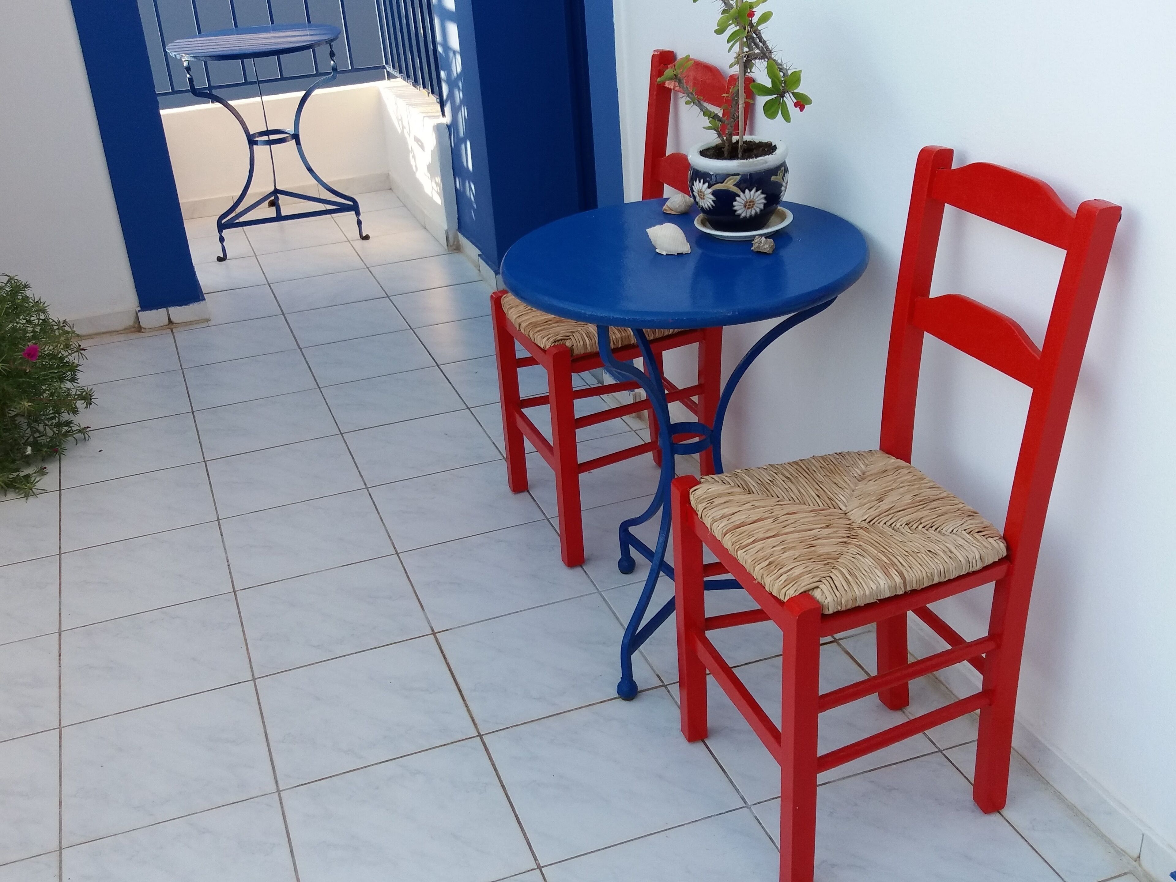 Een klein terras buiten met een blauwe tafel en rode stoelen, een potplant en een blauwe metalen stoel.