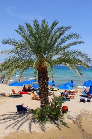A beach with palm trees, blue umbrellas, and sun loungers. - Psaras Apartments (Hersonissos)