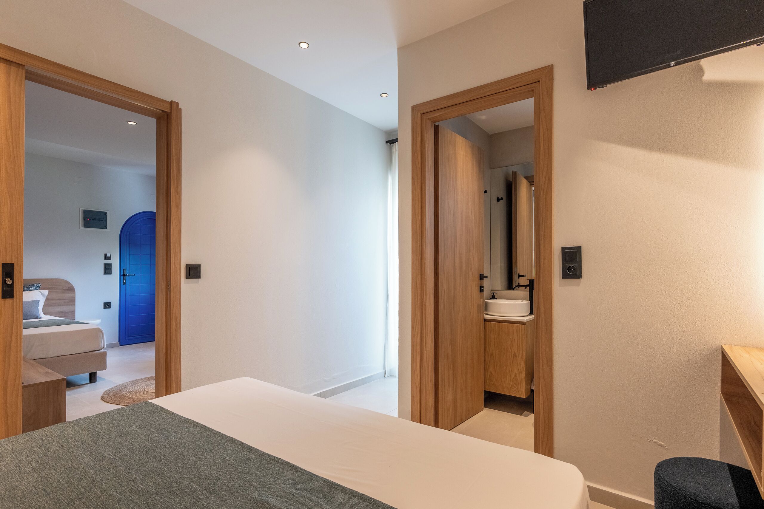 Habitación de hotel moderna con una cama, un escritorio, un baño con lavamanos y una puerta azul al fondo.