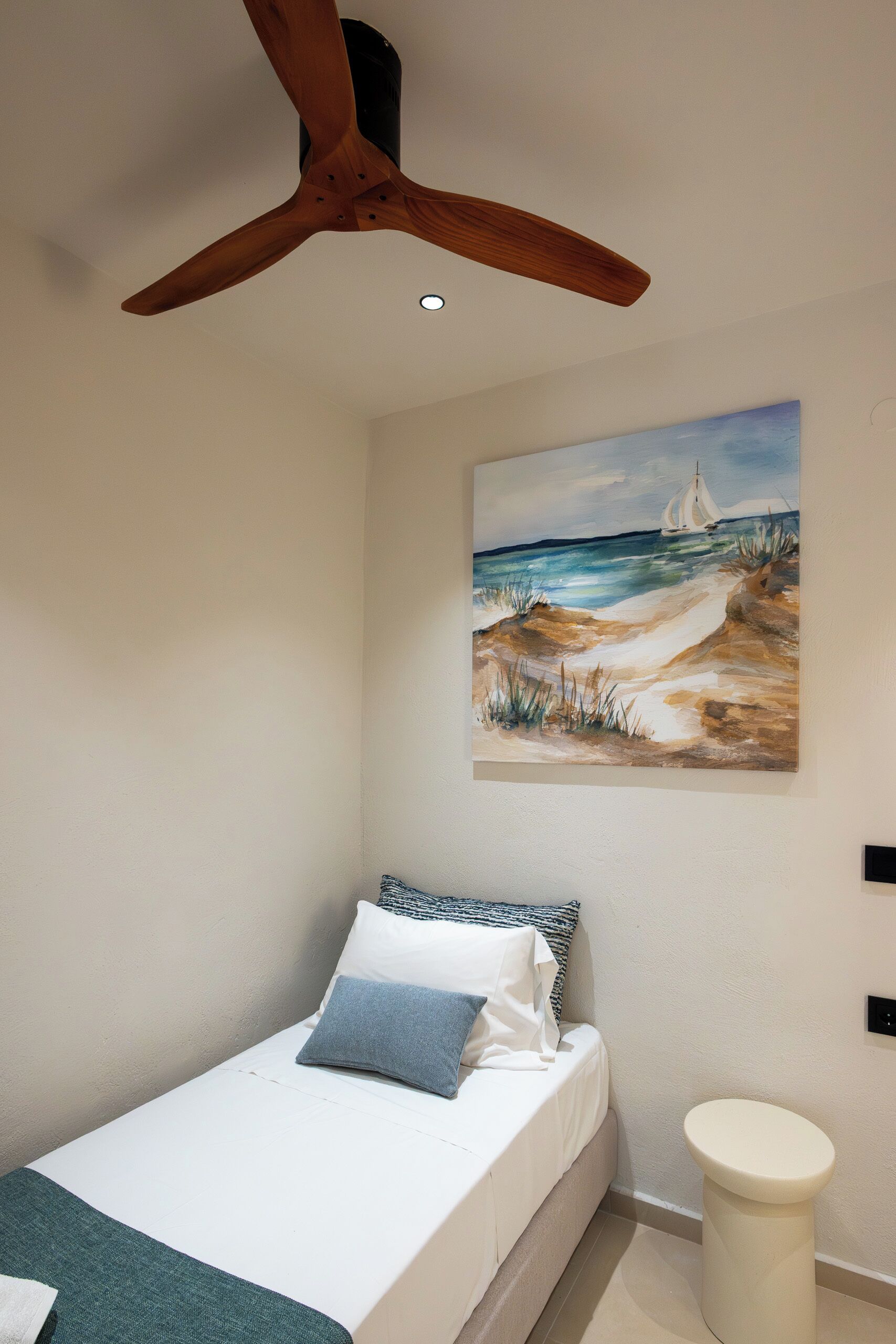 Une chambre à coucher avec un lit, un tableau représentant une scène de plage, un ventilateur de plafond et un petit tabouret blanc.
