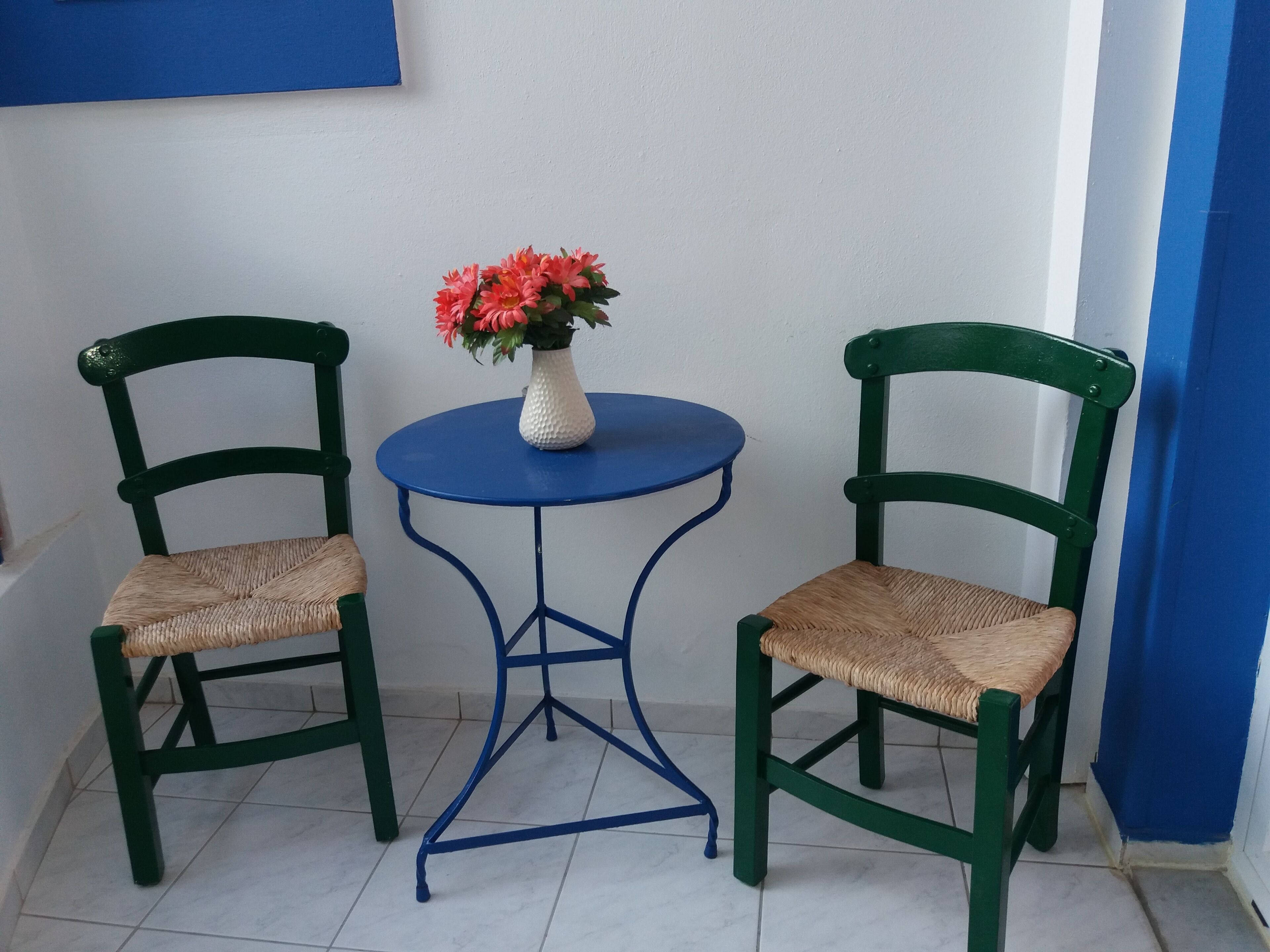 Dos sillas verdes con asientos de mimbre y una pequeña mesa redonda con un jarrón de flores blancas en el centro.