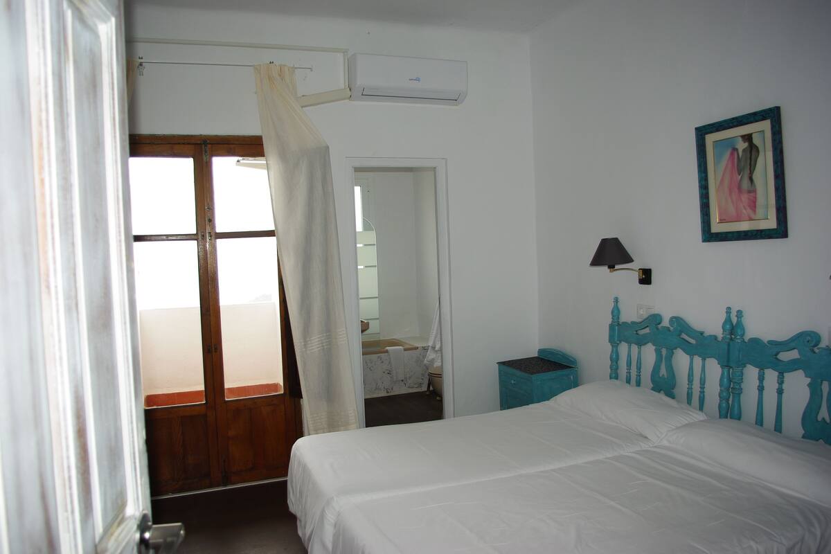 A bedroom with a bed, a nightstand, a wall-mounted lamp, a painting, and a bathroom visible through an open door.