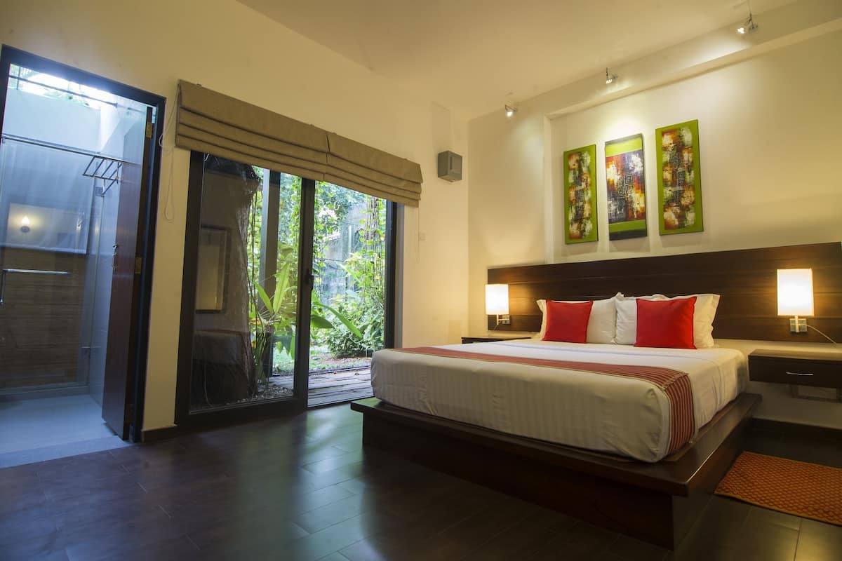 A modern hotel room with a large bed, a sliding glass door leading to a garden, and a nightstand with a lamp.