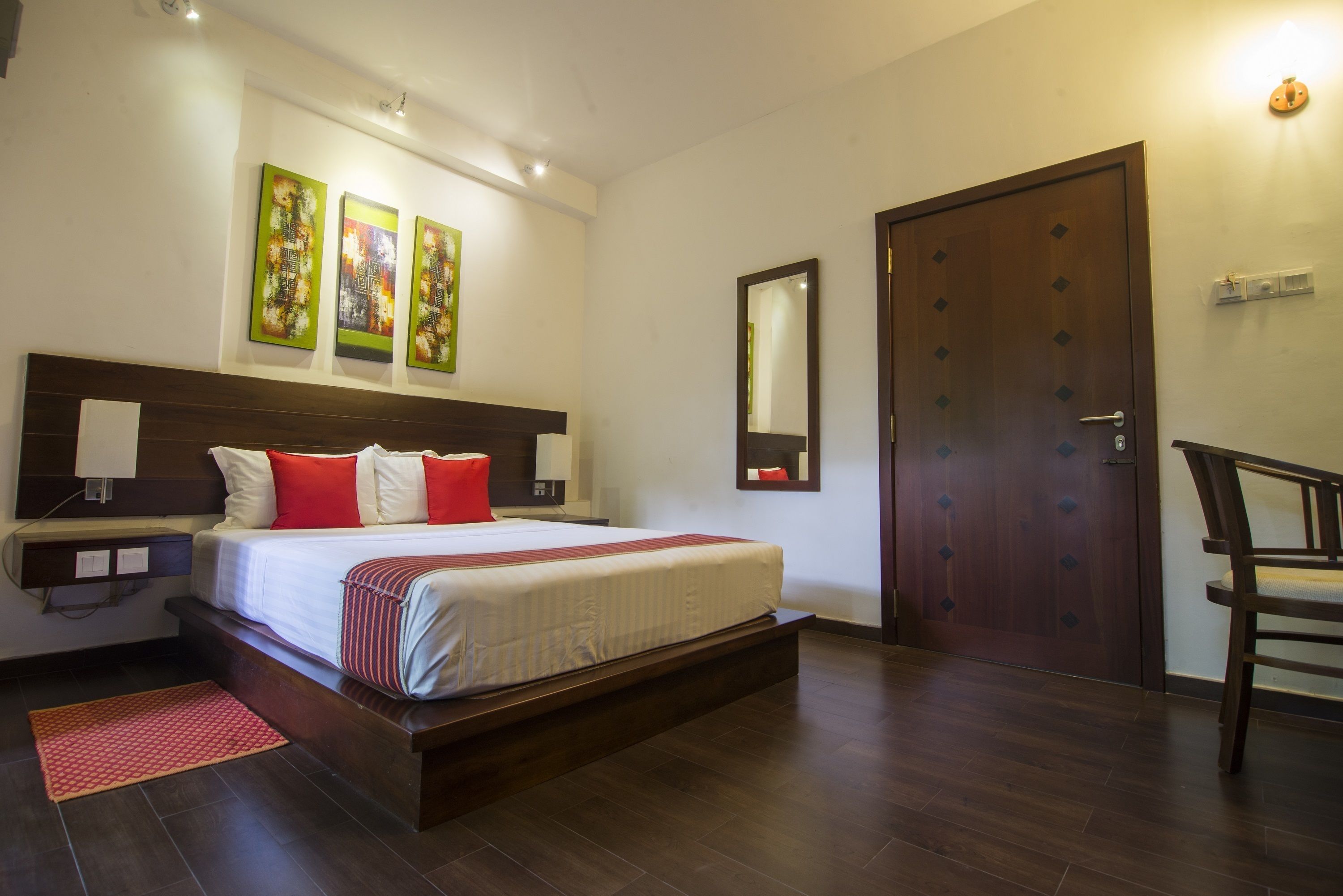 A hotel room with a large bed, a wooden headboard, a red and white striped bedspread, a red pillow, a wooden nightstand with a lamp, a red rug, a wooden door, and a mirror on the wall.