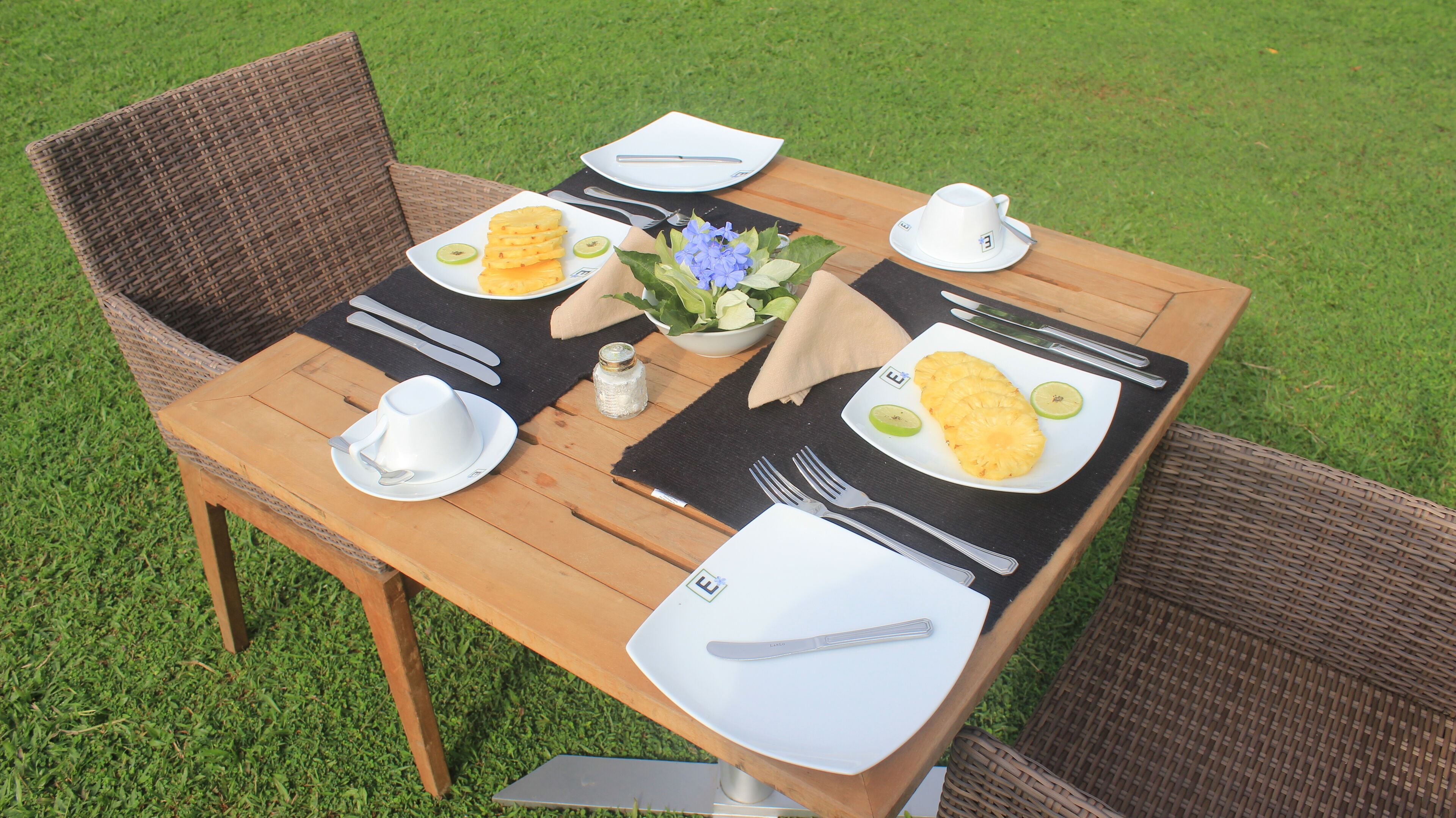 Een houten eettafel buiten gedekt, met borden eten, bestek en een kleine vaas met bloemen.