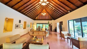 A spacious living area with a high wooden ceiling, a dining table with chairs, a sofa, and a large window offering a view of the outdoors. - Villa Escondite (Sri Jayawardenepura Kotte)