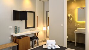 Une chambre d’hôtel moderne équipée d’une télévision à écran plat, d’une coiffeuse avec miroir, d’une table de chevet avec une lampe et d’un lit soigneusement fait avec des serviettes.