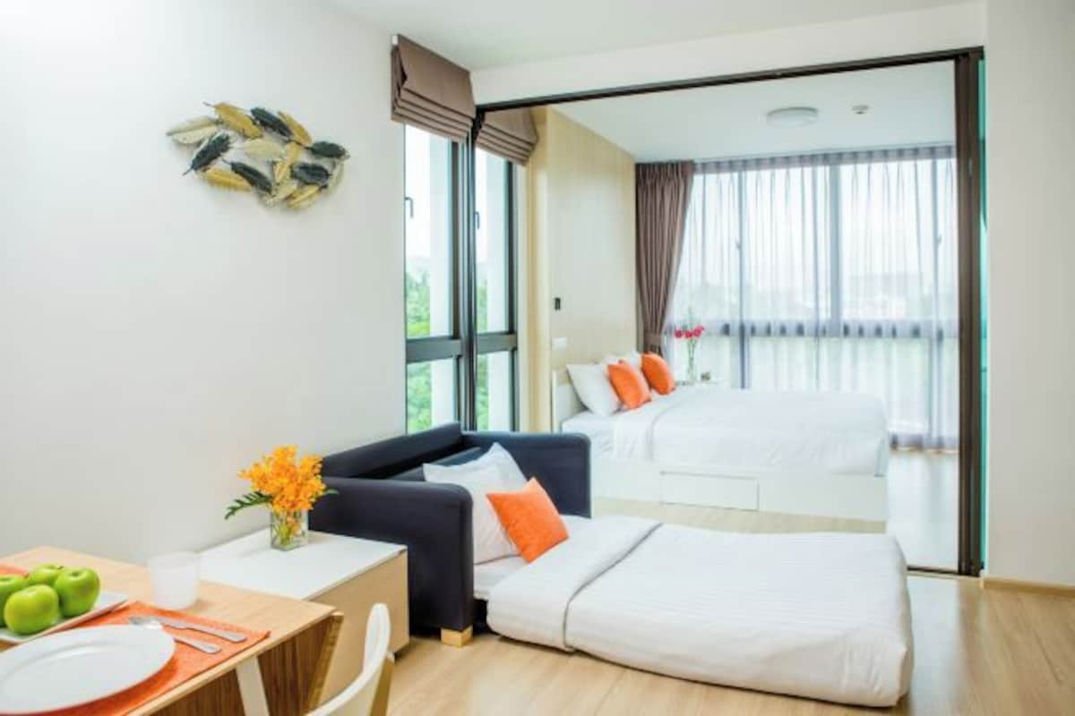 A modern hotel room with a sofa, a dining table with apples, a vase of flowers, and a bed with orange pillows.