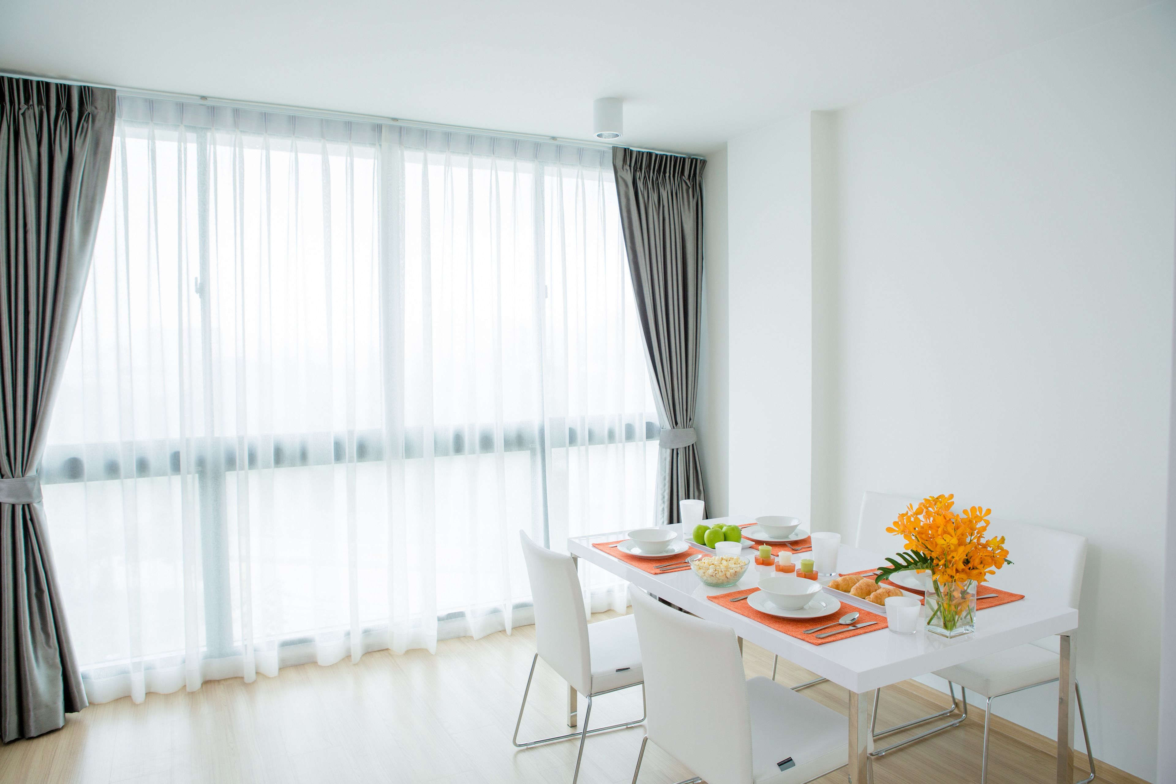 Un comedor moderno con una mesa blanca dispuesta para dos personas, con manteles individuales naranja, sillas blancas y un jarrón con flores naranja.
