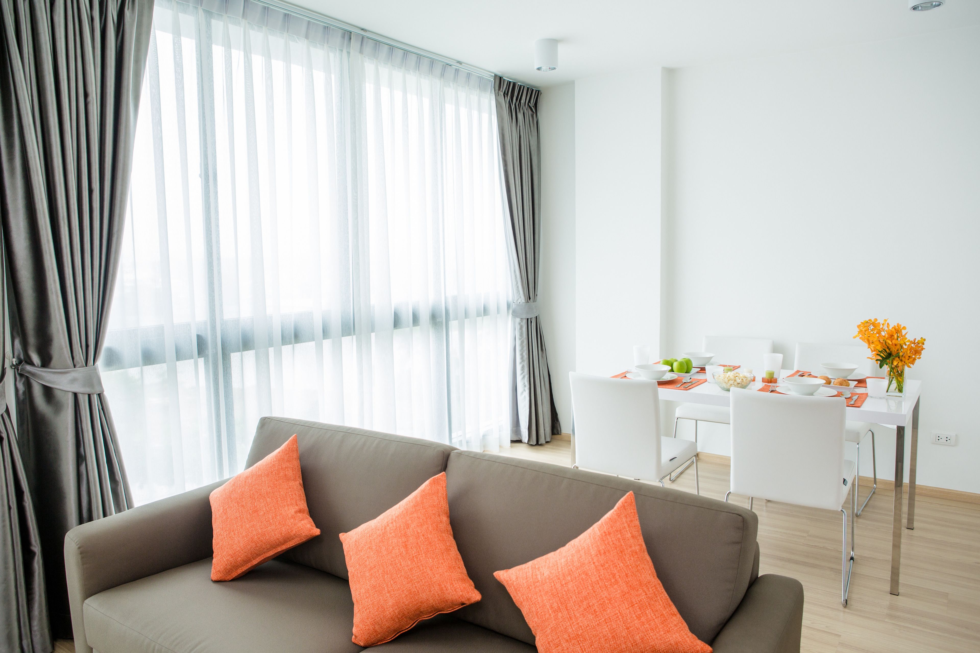 A modern living room with a grey sofa, orange cushions, a dining table with white chairs, and a large window with sheer curtains.