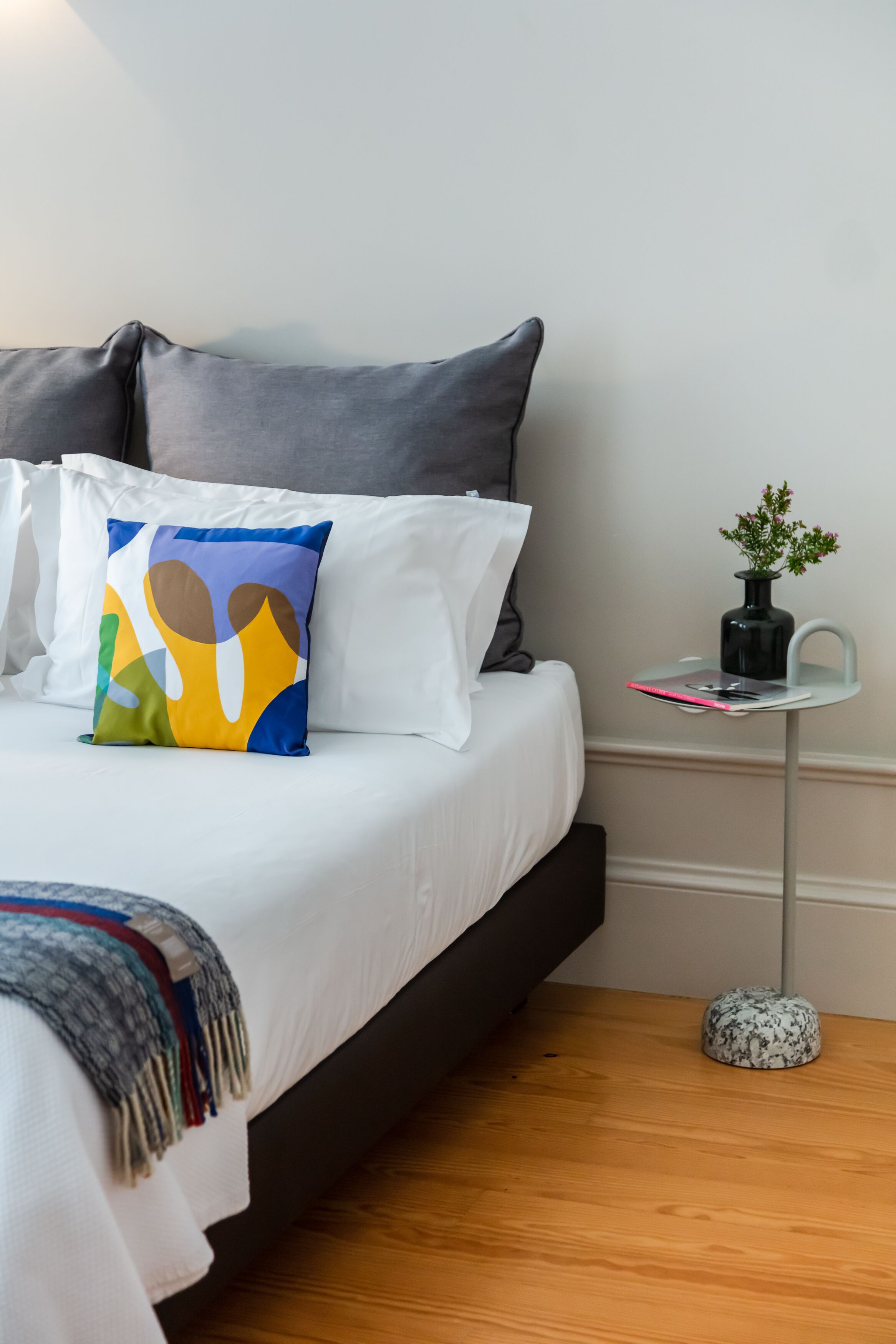 A neatly made bed with white bedding and a colorful abstract pillow, a small side table with a vase, and a wooden floor.