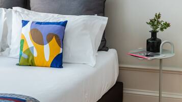 A neatly made bed with white bedding and a colorful abstract pillow, a small side table with a vase, and a wooden floor.