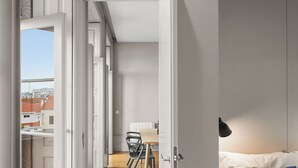 A modern hotel room with a wooden floor, a white bed with a colorful pillow, and a view of the city through an open door. - Baumhaus Serviced Living - Art & Design Apartments (Porto)