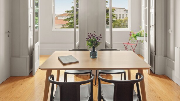 A dining area with a wooden table and chairs, a vase with flowers, and large windows with a view of buildings and trees. - Baumhaus Serviced Living - Art & Design Apartments (Porto)