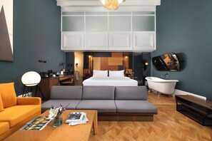 A modern hotel room with a sofa, a coffee table, a TV, a bathtub, and a desk with a lamp.