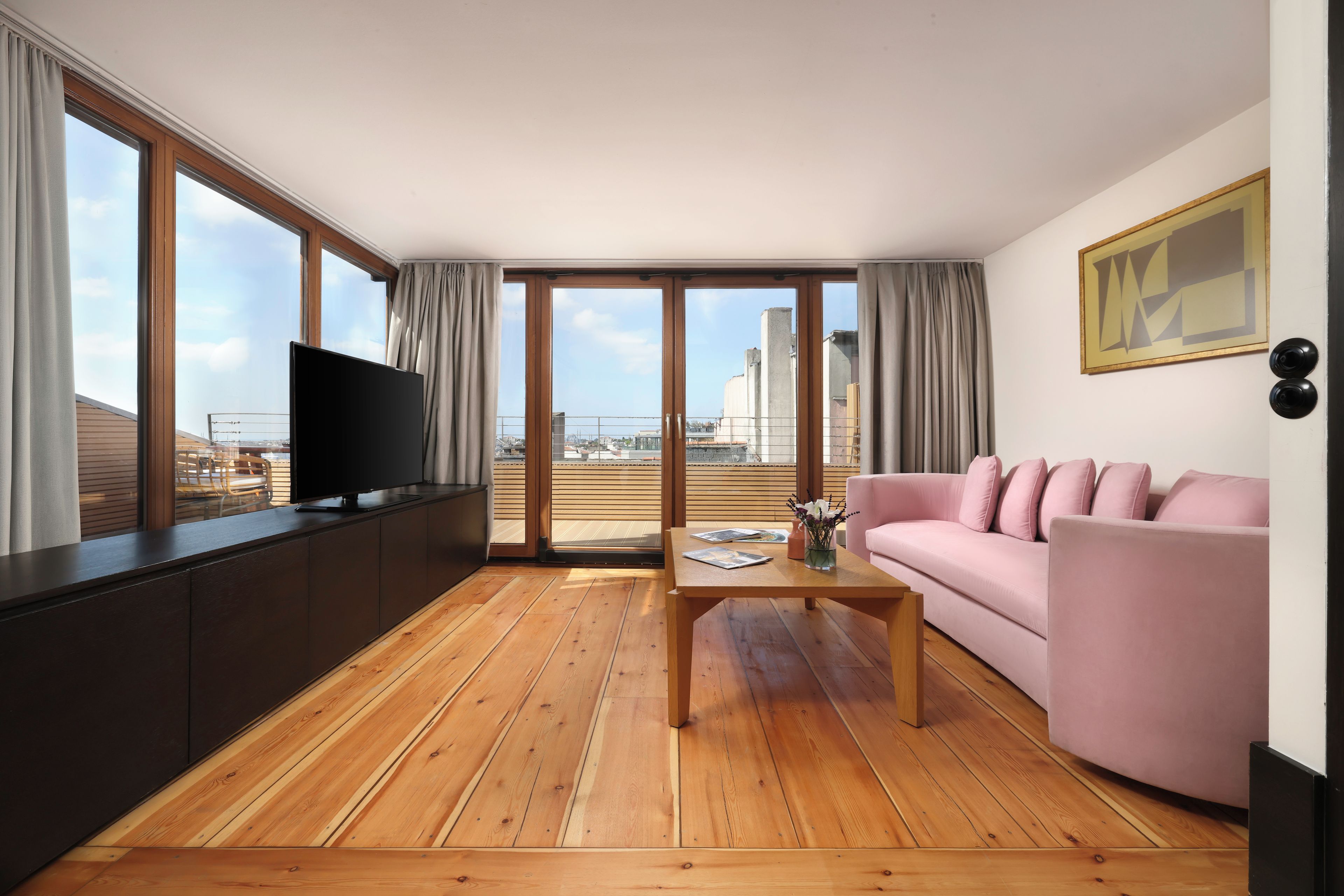 Ein modernes Wohnzimmer mit Fernseher, einem rosafarbenen Sofa, Holzböden und großen Fenstern mit Blick auf die Stadt.