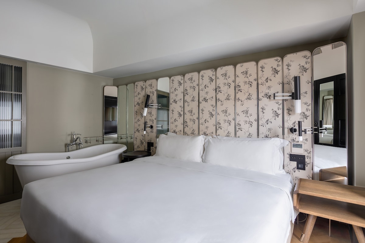 Une chambre d’hôtel moderne avec un grand lit, une baignoire, un banc en bois et un miroir agrémenté d’un papier peint à motifs floraux.