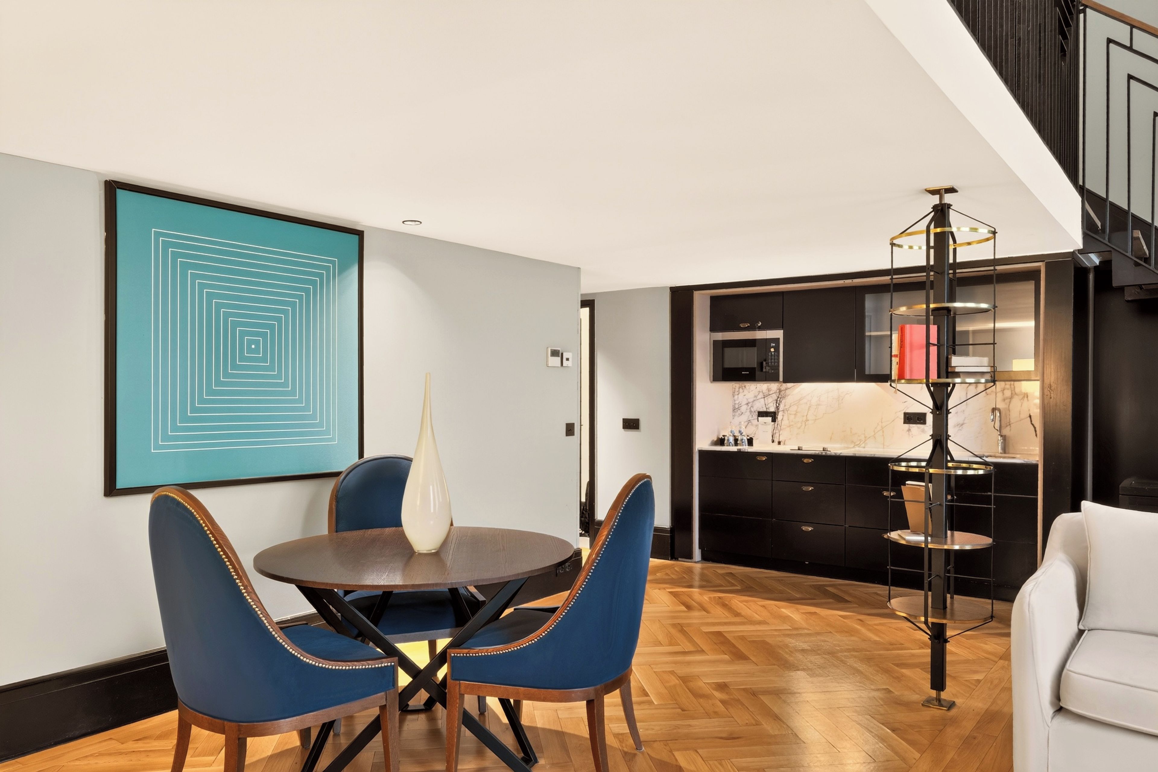 A modern dining area with a round table and blue upholstered chairs, a kitchen with black cabinetry, and a staircase with a black railing.