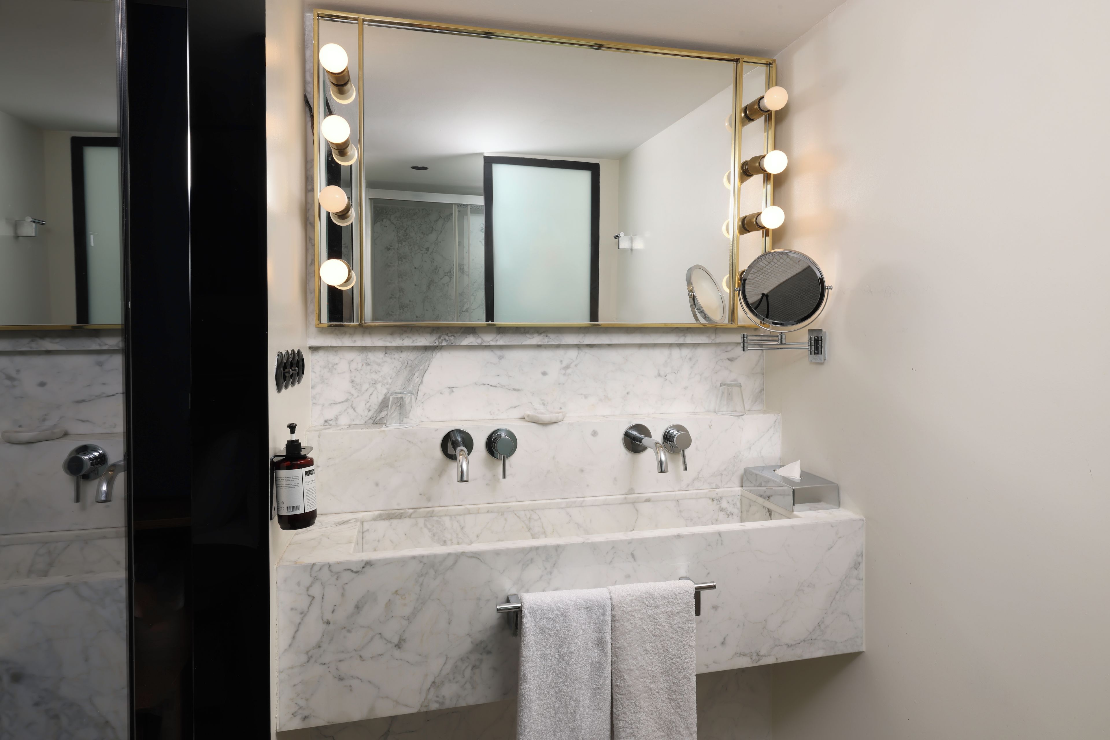 Une salle de bain avec un meuble-lavabo en marbre, un grand miroir avec éclairage intégré, un miroir rond et un porte-serviettes.