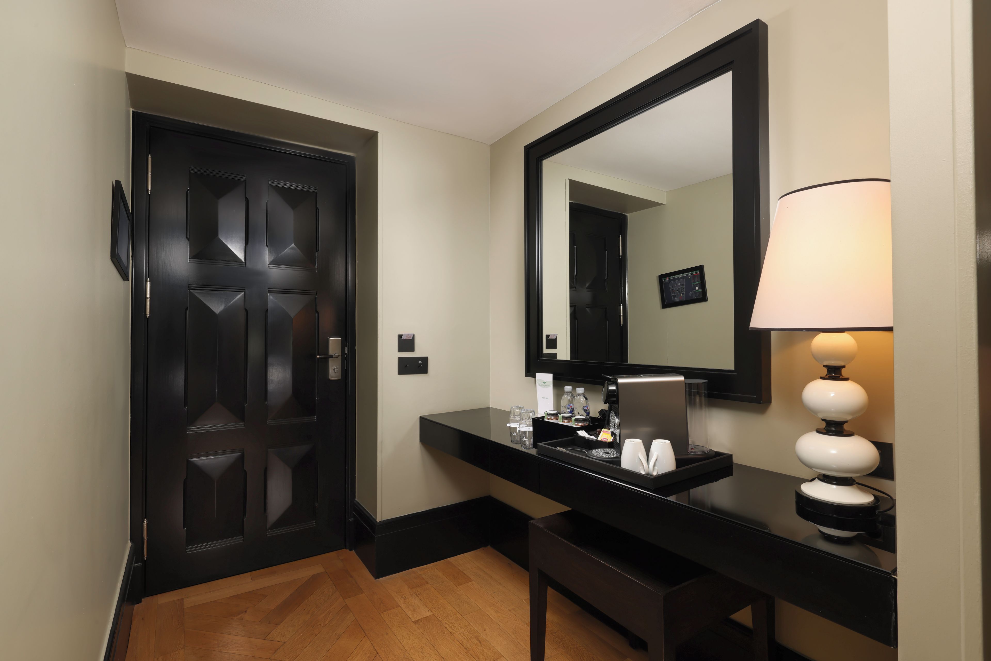 Ein Hotelzimmer mit einer dunkelholznen Tür, einer schwarzen Arbeitsplatte mit einem Spiegel, einer Lampe und einem kleinen Tisch mit verschiedenen Gegenständen.