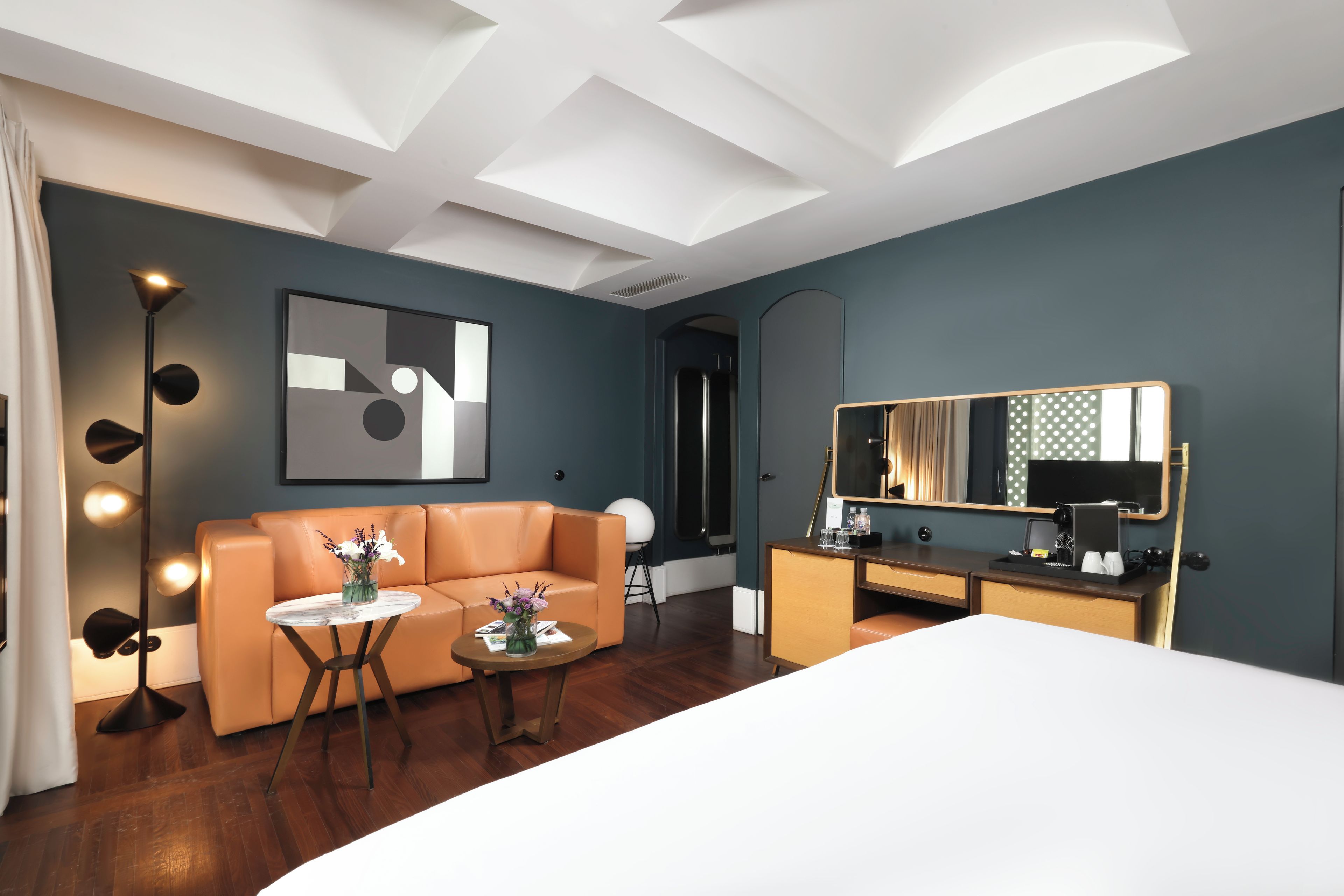 Ein modernes Hotelzimmer mit einer Couch, einem runden Tisch mit Blumenvase, einem Bett, einem Schreibtisch mit Spiegel und einem großen abstrakten Gemälde an der Wand.
