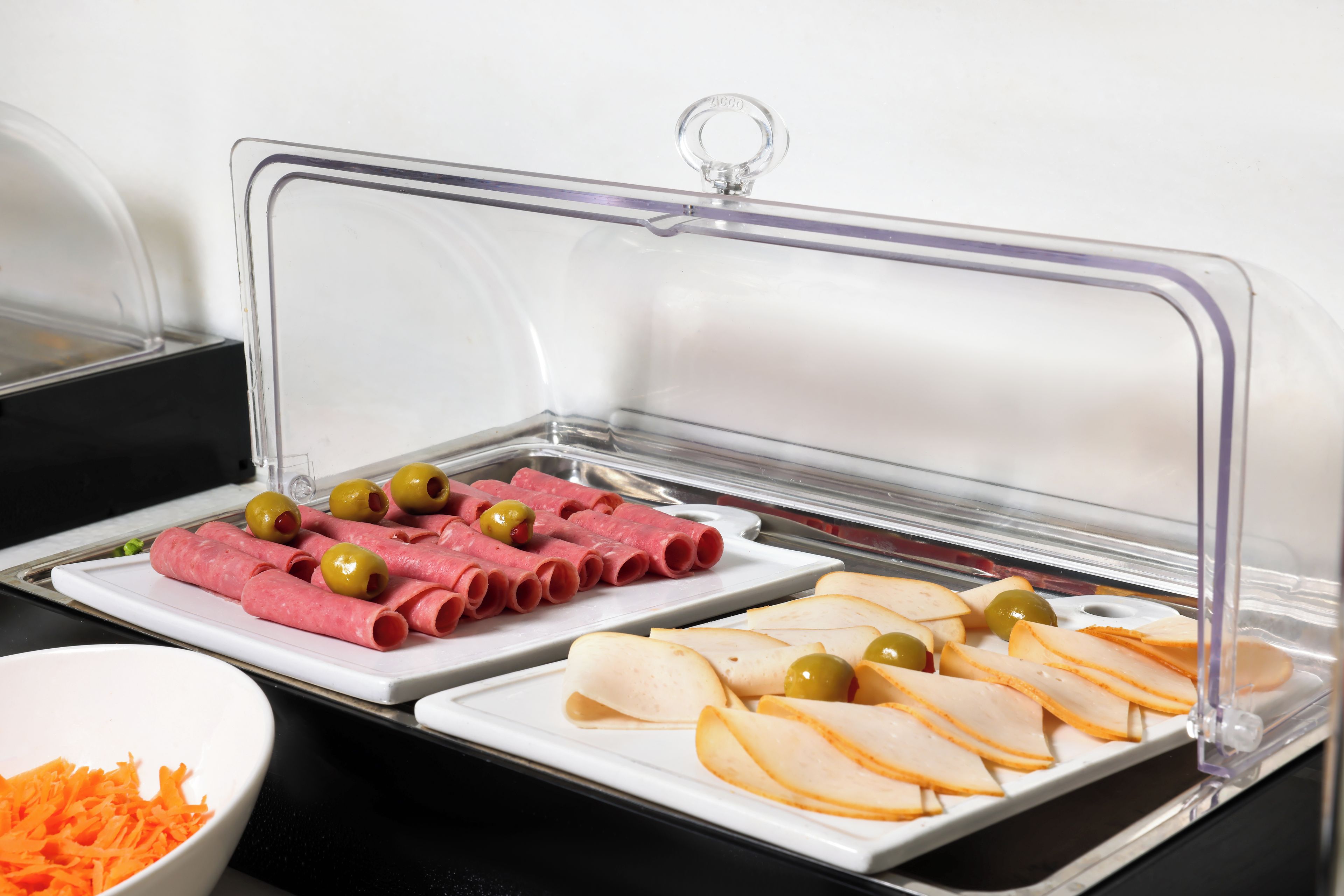 Buffet com diversos alimentos, incluindo carnes fatiadas e queijos, coberto por uma cúpula de plástico transparente.