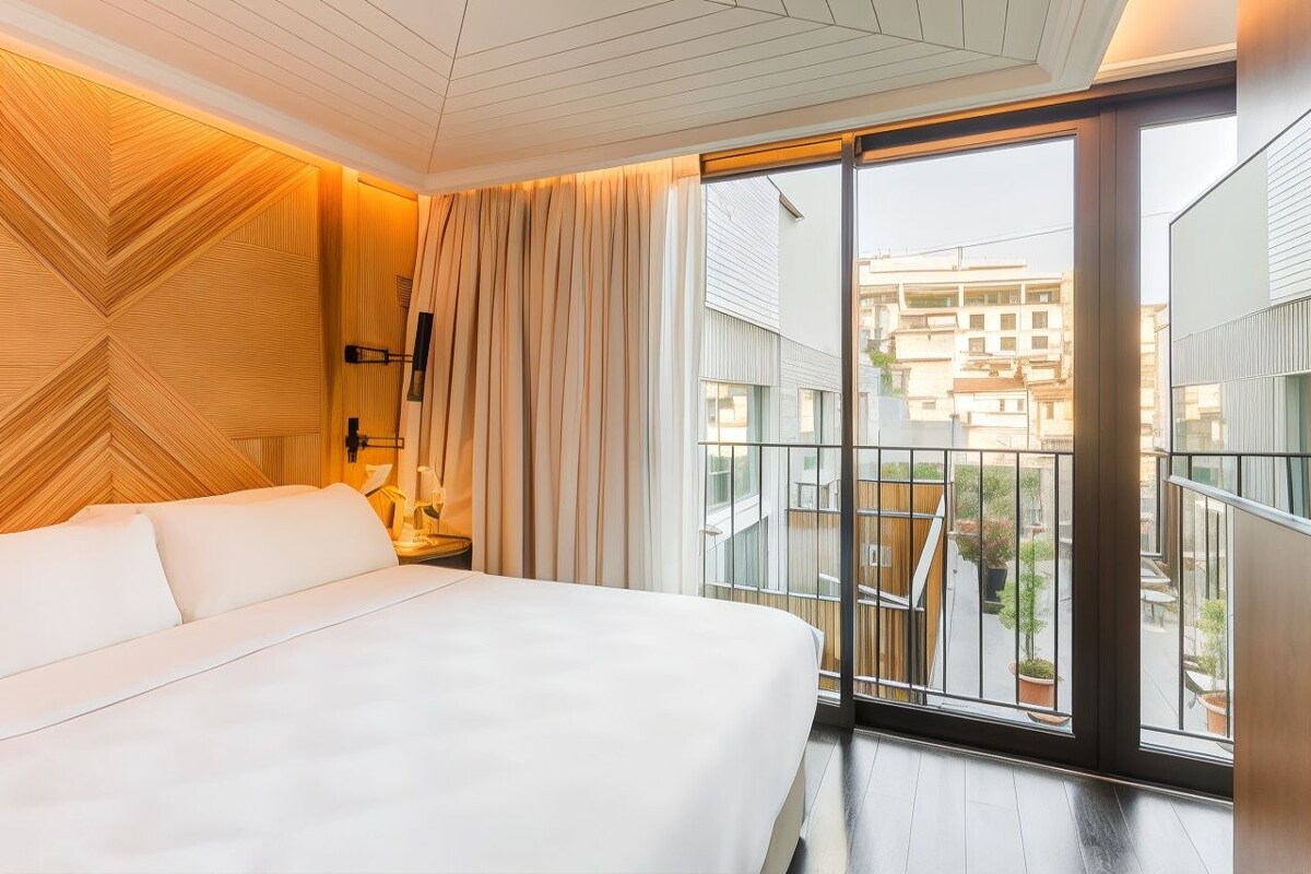 A hotel room with a bed, a nightstand, a lamp, a large window with a view of buildings, and a wooden wall panel.