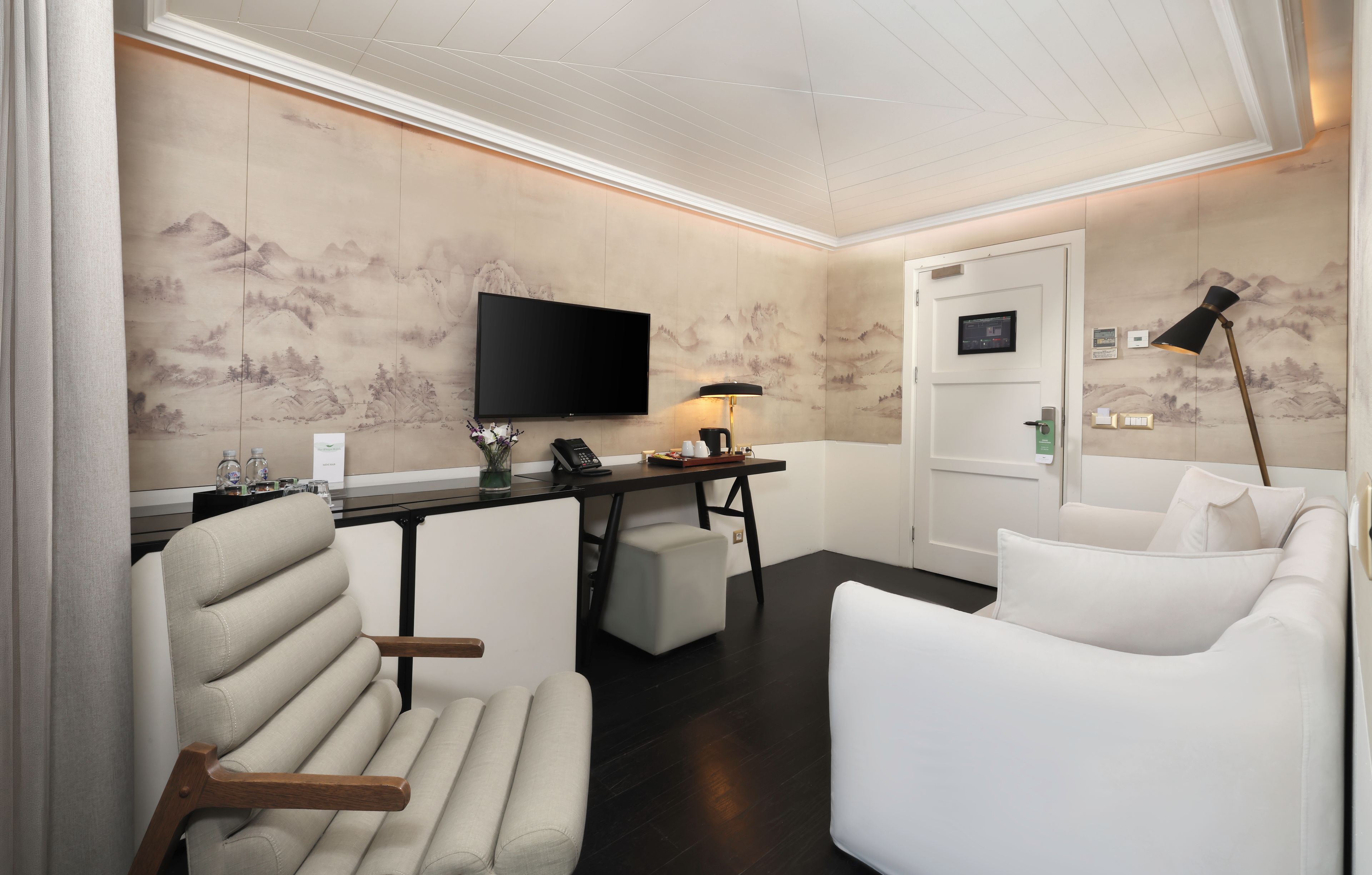 Habitación de hotel moderna con televisión de pantalla plana montada en la pared, un escritorio con lámpara y dos sofás blancos.