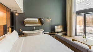 Een moderne hotelkamer met een groot bed, een badkuip, een zithoek met een tafel, en een balkon met tuinmeubilair.