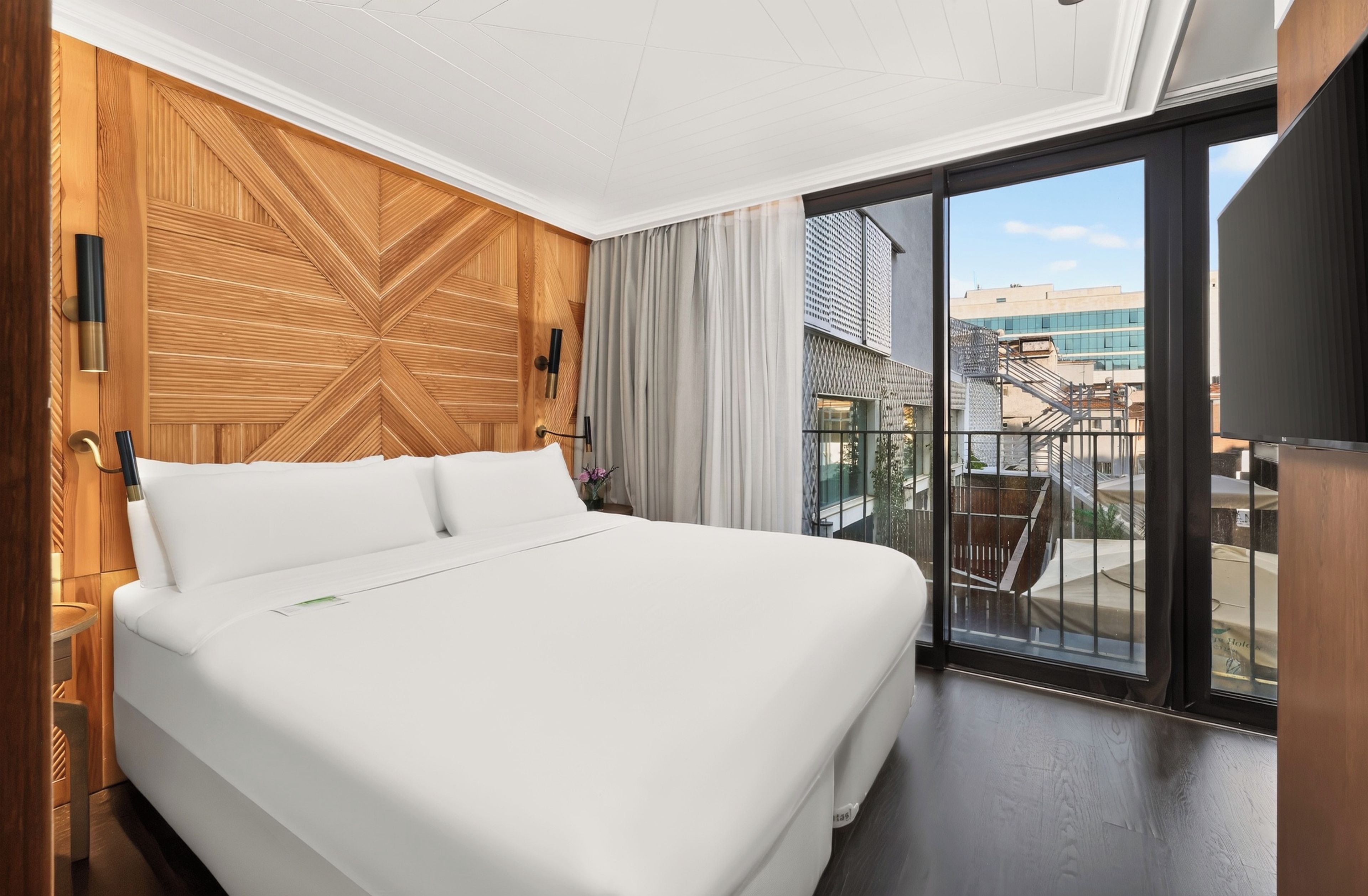 A hotel room with a large bed, wooden panel wall, a balcony with a view of buildings, and a television.