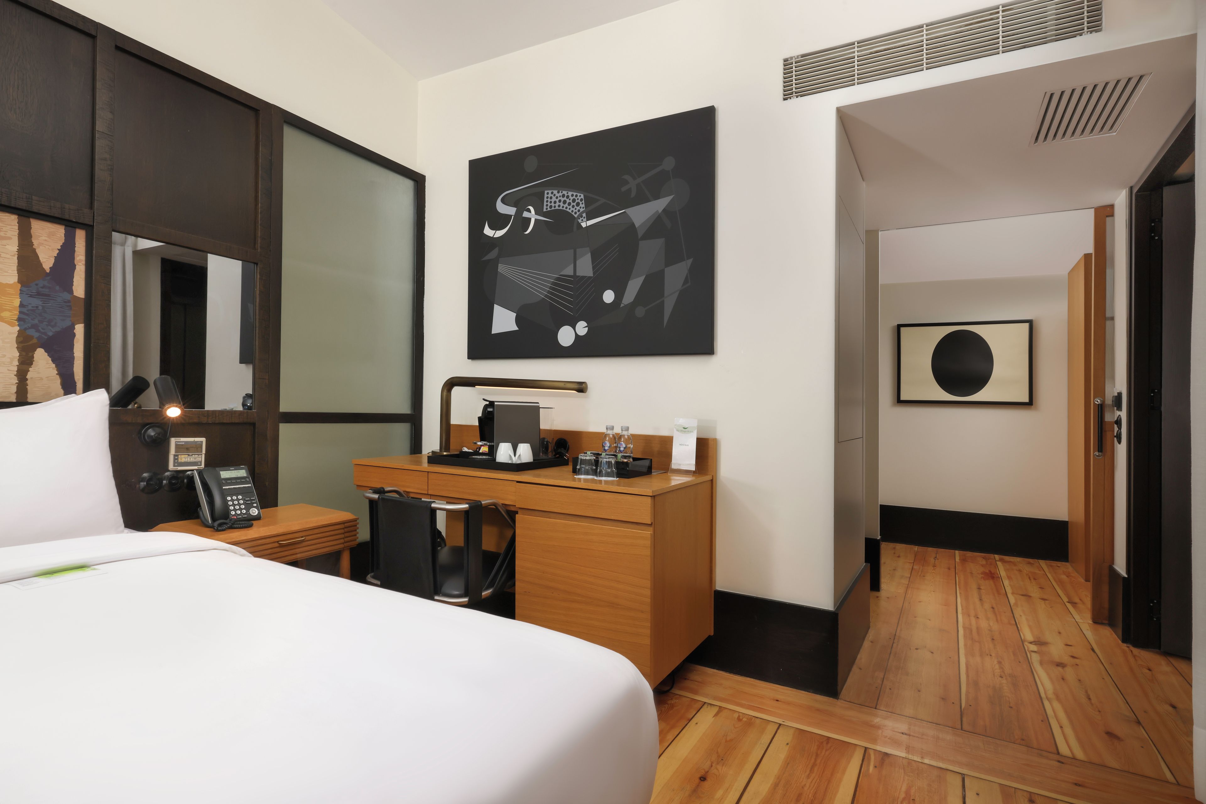 A hotel room with a bed, a desk with a telephone, a coffee machine, and a painting on the wall.